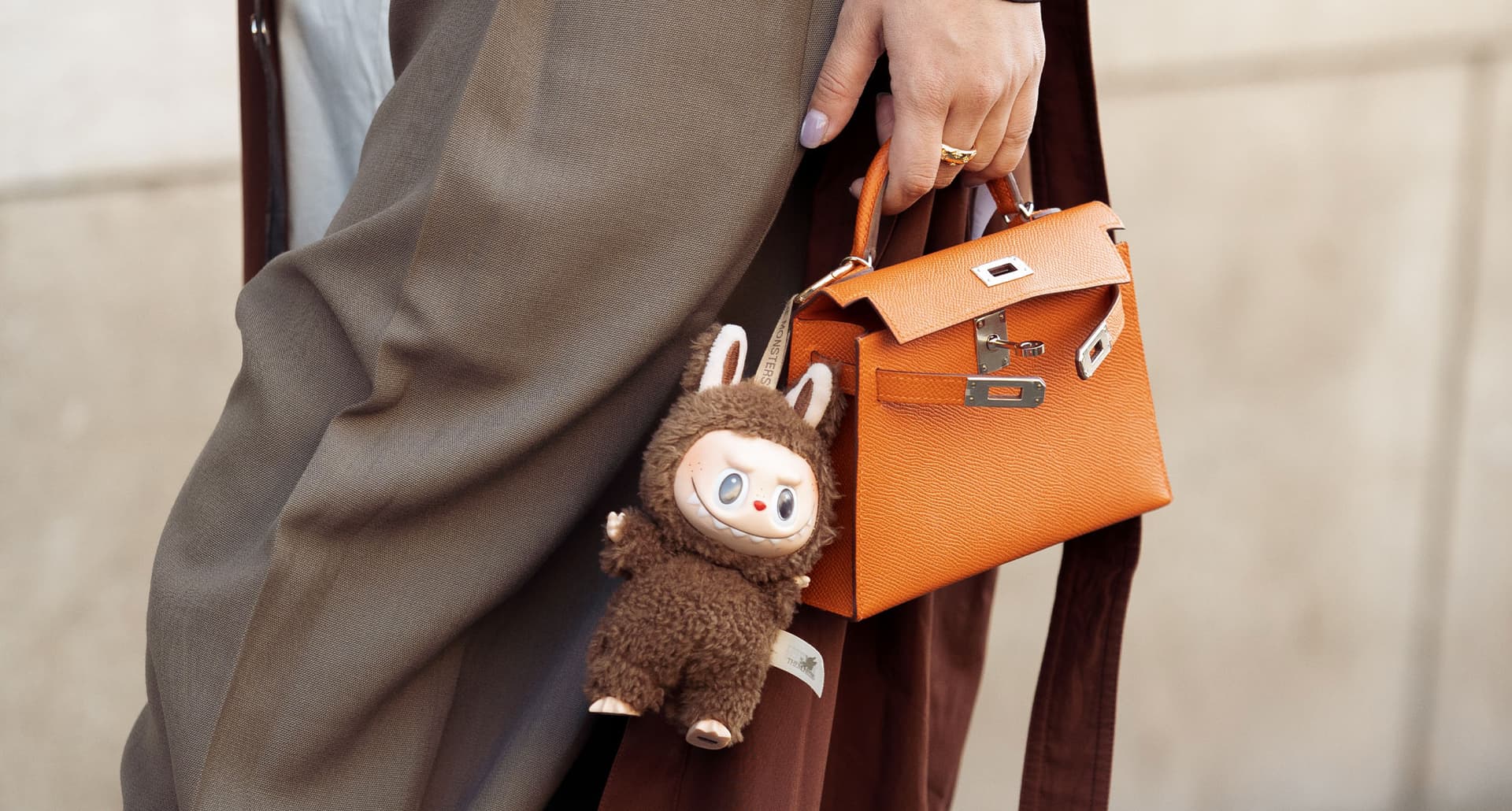 labubu-bag-charm-trend: A close up photograph of a woman in brown pants and a terracotta colored coat carries a bright orange Birkin bag with a chocolate brown LABUBU keychain on it.